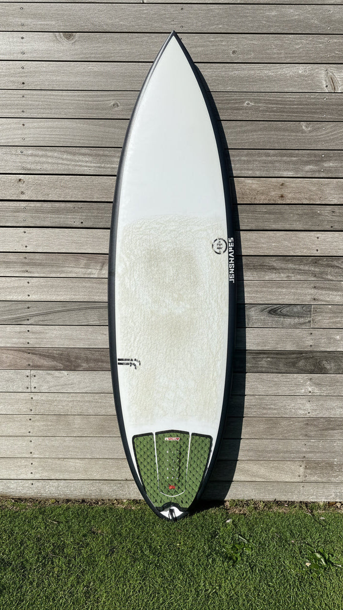 A white surfboard with a black edge and a green leash attachment patch, photographed from above on a weathered wooden deck next to grass.