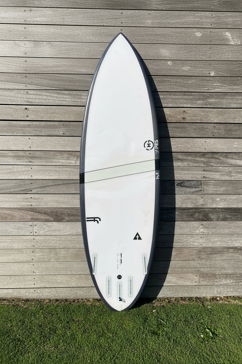 A white surfboard with a black border, photographed from above on a weathered wooden deck with grass visible on the right side.